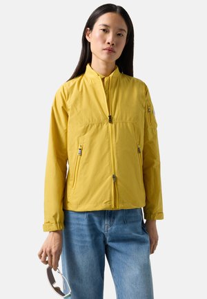 Młoda kobieta w jaskrawo żółtej kurtce na zamek i niebieskich dżinsach, trzymająca przeciwsłoneczne okulary w jednej ręce na jednolitym tle.