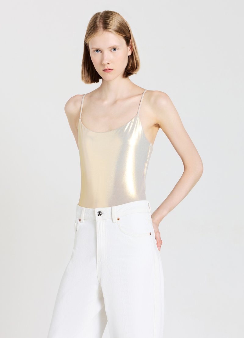Body metallico dorato con spalline sottili abbinato a jeans bianchi a vita alta, con dettagli di bottoni e tasche. Texture liscia, design riflettente.