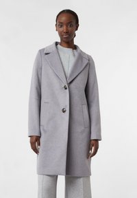 Femme portant un manteau en laine gris clair avec de larges revers et deux boutons, associé à un tricot et un pantalon gris assortis, le tout sur un fond uni.