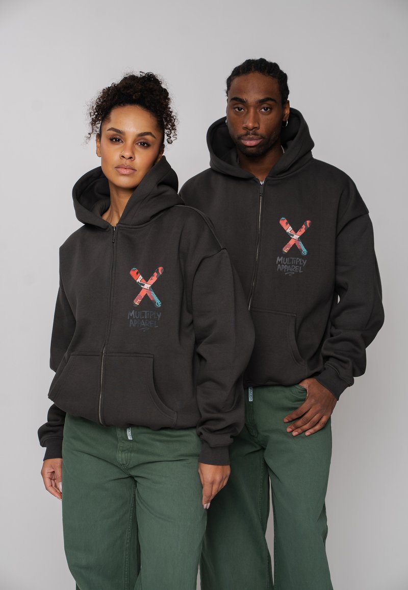 Două persoane purtând hanorace negre cu fermoar cu logo-ul colorat "Multiply Apparel" stau pe fundal gri, ambele purtând pantaloni verzi.