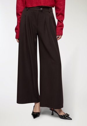 Donna che indossa pantaloni a gamba larga marroni scuri, un maglione rosso e tacchi neri a punta con decorazioni a fiocco e borchie.