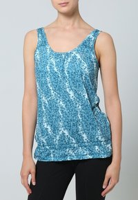 Kvinna i ärmlös blå t-shirt med geometriskt mönster och svarta leggings, som står mot en enkel ljus bakgrund.