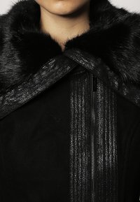 Collar de piel sintética negra en una chaqueta negra, que presenta un panel vertical texturizado y brillante, y un cierre de cremallera oculto. Primer plano de la prenda superior.