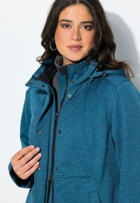 Nainen, jolla on tummat hiukset, käyttää korvakoruja ja sinistä vetskarijakkua, mukauttaen etuosaa kaulusta vasten valkoista taustaa vasten.