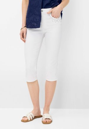 Femme portant un pantalon court blanc avec des détails cloutés sur la poche avant, un haut bleu marine et des sandales blanches à enfiler, debout sur fond blanc.