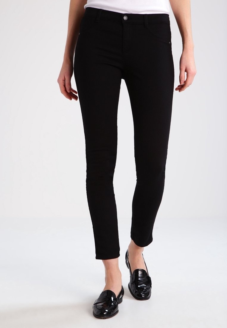 Femme portant un pantalon noir slim avec des poches avant et des mocassins noirs brillants, debout devant un fond clair uni.