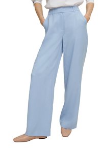 Femme portant un pantalon large bleu clair et des ballerines beiges, les mains dans les poches, vue de la taille jusqu'aux pieds sur fond blanc.