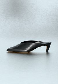 Γυναικείο mule από μαύρο δέρμα με μυτερή μύτη και λεπτό τακούνι μεσαίου ύψους, πάνω σε ανοιχτό γκρι επιφάνεια.