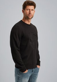 Sudadera negra de cuello redondo hecha de una tela suave, con un diseño de logo texturizado en la parte delantera y puños y dobladillo acanalados. Combinada con jeans azules.