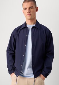 Navy blue jacket made of smooth fabric, featuring a snap button closure, elastic cuffs, and a simplified collar design. Worn over a light blue shirt.