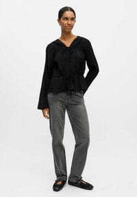 Zwarte geplooide blouse met een V-hals en een koordtaille, gecombineerd met grijze rechte jeans en zwarte platte schoenen. Glad materiaal, relaxte pasvorm.
