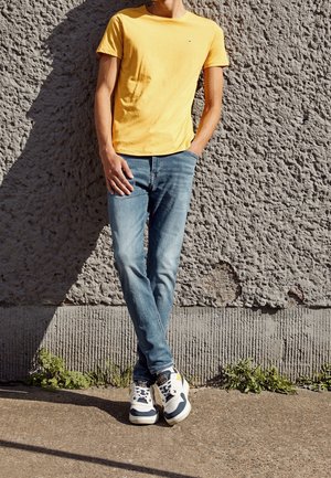 Żółty bawełniany t-shirt, niebieskie dżinsy i białe tenisówki z granatowo-żółtymi akcentami, stojące na tle teksturowanej szarej ściany.