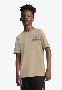 Μπεζ βαμβακερό μπλουζάκι με κοντά μανίκια, στρογγυλή λαιμόκοψη και τυπωμένο γραφικό λογότυπο στο στήθος. Συνδυασμένο με μαύρο παντελόνι.