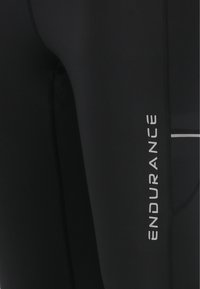 Zwarte sportleggings van soepel, rekbaar materiaal. Voorzien van witte "ENDURANCE" letters en reflecterende accenten langs de naden.