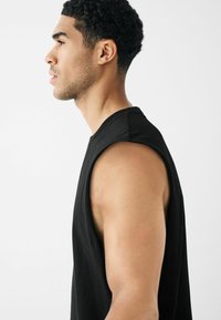 Haut sans manches noir en tissu doux, avec une coupe décontractée et des emmanchures ouvertes, affichant des coutures latérales et une texture lisse.