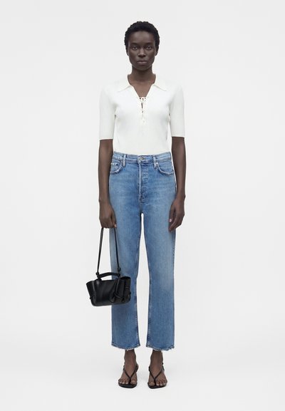 Γυναίκα με λευκή μπλούζα με κοντά μανίκια, ψηλόμεσα μπλε τζιν, μαύρη τσάντα και μαύρα σανδάλια με τακούνι στέκεται μπροστά σε απλό φόντο.