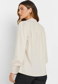 Blouse beige à manches longues avec une coupe décontractée, présentant un dos plissé et des poignets smockés, fabriquée en tissu léger. Texture lisse.