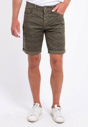 Homme portant un short vert olive retroussé avec un motif discret de feuilles et des baskets blanches, debout sur fond blanc.