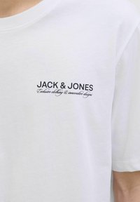 Valkoinen puuvillainen T-paita, jossa on pyöreä kaula-aukko, lyhyet hihat ja vasemmassa rintapielessä mustalla painettuna teksti "JACK & JONES."