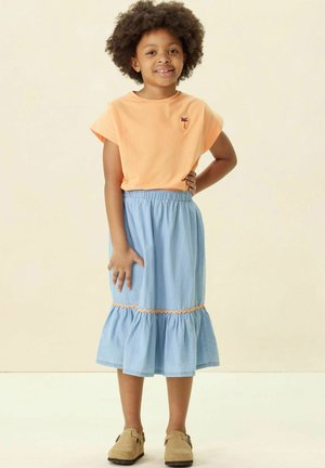 T-shirt court à manches courtes de couleur orange clair avec un petit motif de palmiers, associé à une jupe en denim bleu avec un ourlet volanté et des bordures orange. Chaussures beiges.