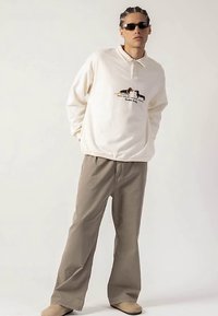 Krémové polokošeľa s dlhým rukávom, golierom a grafickou potlačou dvoch psov, ktoré sú skombinované s béžovými, voľnými nohavicami a svetlohnedou obuvou.