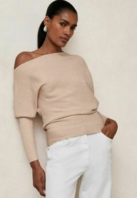 Beige gerippter Off-Shoulder-Pullover mit langen Ärmeln, kombiniert mit weißen Hosen. Auffällige Textur und Passform betonen lässigen Komfort.