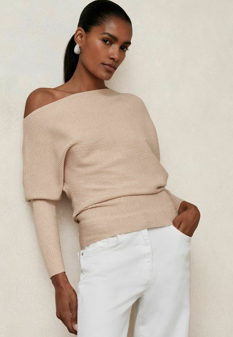 Beige gerippter Off-Shoulder-Pullover mit langen Ärmeln, kombiniert mit weißen Hosen. Auffällige Textur und Passform betonen lässigen Komfort.