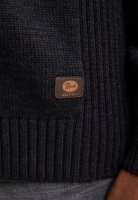 Dunkel gestrickter Pullover mit strukturiertem Rippmuster und einem braunen Lederschild mit der Aufschrift "Petrol Industries" an der unteren Vorderseite.
