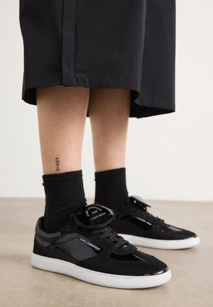 Piernas con calcetines negros y zapatillas negras Karl Lagerfeld con suelas blancas, de pie sobre un suelo beige, con una falda negra visible arriba.