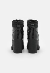 Botas de tobillo negras con tacón, acabado de cuero brillante, puño texturizado suave y tacones robustos en bloque. Presenta detalles en forma de lazo en la parte posterior.