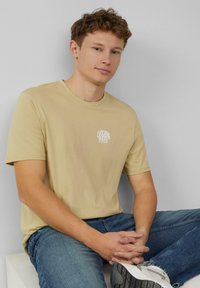 Lichtbeige katoenen t-shirt met een klein wit grafisch tekstlogo, gecombineerd met blauwe spijkerbroek en grijze schoenen. Gladde textuur, ronde halsontwerp.