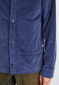 Navy corduroy jacket with a buttoned front, two lower pockets, and long sleeves. Soft texture with a casual fit.