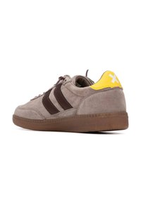 Zapatillas de ante en gris claro con rayas marrones, acento en el talón amarillo y suela marrón texturizada. Presenta puntera redondeada y diseño de cordones.