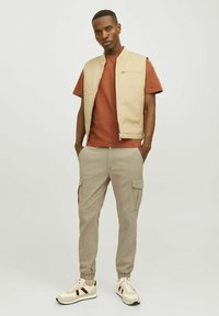 Beige sleeveless vest over a rust t-shirt, paired with light brown cargo pants and cream sneakers with maroon accents.