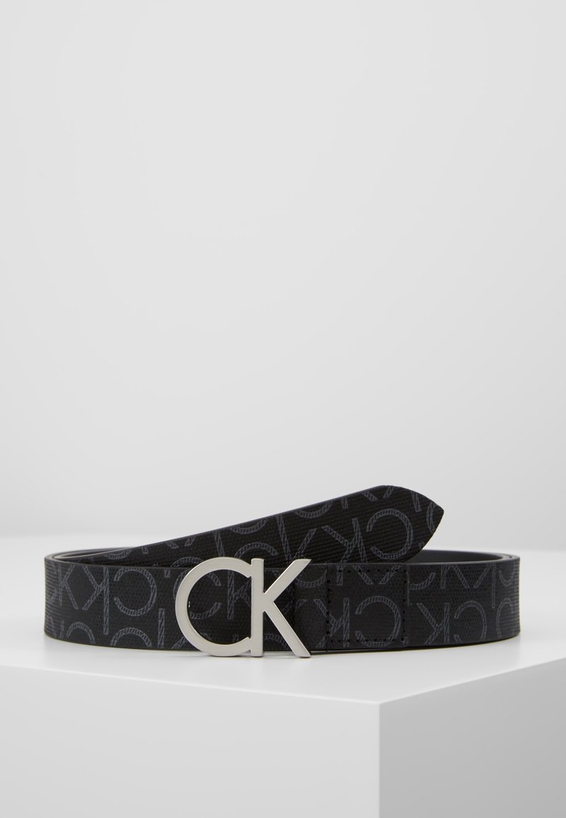 Ceinture en cuir noire avec motif répétitif "CK" et boucle logo "CK" argentée présentée sur une surface blanche avec un fond gris.
