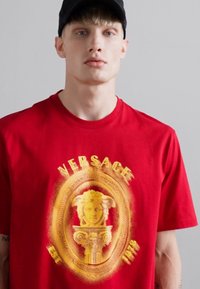 Jaunas vyras, vilkintis juodą kepurę ir raudoną Versace marškinėlį su auksiniu Medūzos logotipu ir tekstu "EST 1978" ant vienspalvio šviesaus fono.