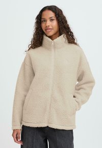 Beežist fliisist jope kõrge krae, esikinnituse ja küljetaskutega. Omadusteks on tekstureeritud, pehme pind ja lõdvestunud lõige.