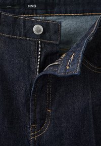 Jean en denim bleu foncé avec une fermeture éclair ouverte, un bouton visible et des surpiqûres orange contrastantes à la ceinture et à la braguette.