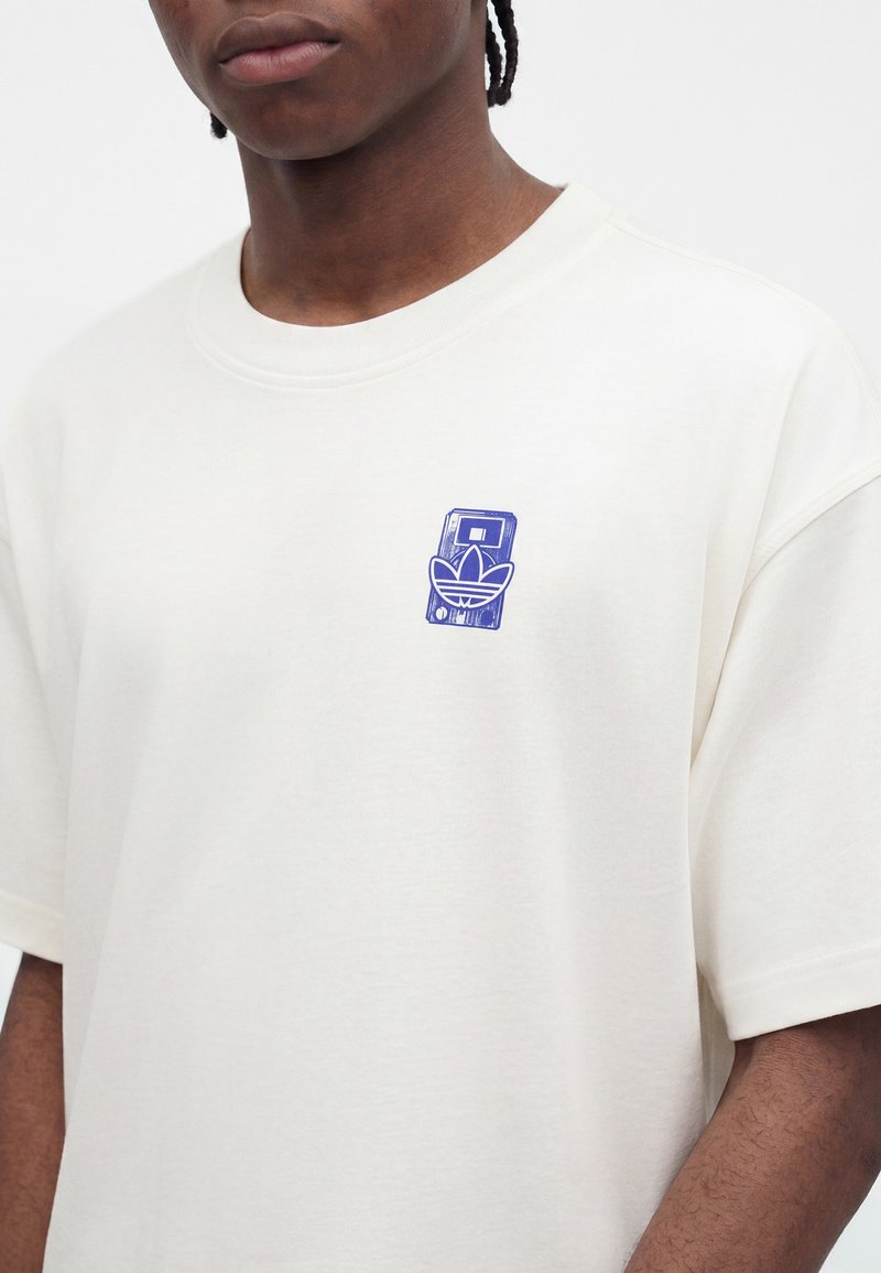 Jeune personne portant un t-shirt blanc avec un petit graphisme bleu combinant une cassette et le logo trefoil d'Adidas sur la poitrine.
