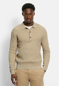 Beige Strickpullover mit kragenförmigem Halsausschnitt, dreiknöpfiger Knopfleiste, gerippten Ärmeln und strukturiertem Stoff. Getragen über einem weißen Hemd.