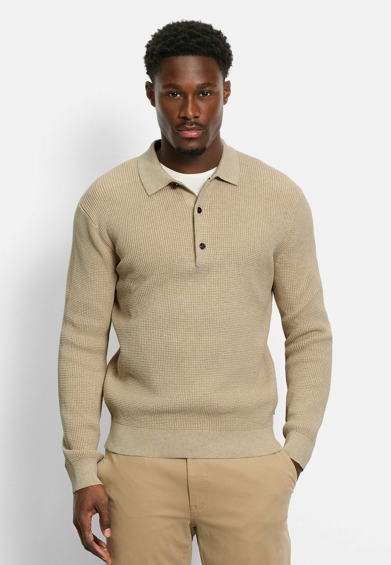 Beige Strickpullover mit kragenförmigem Halsausschnitt, dreiknöpfiger Knopfleiste, gerippten Ärmeln und strukturiertem Stoff. Getragen über einem weißen Hemd.