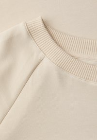 Tela de sudadera beige con cuello acanalado y costuras en los hombros, que presenta una textura suave y una superficie ligeramente texturizada.