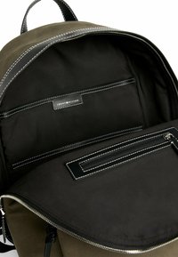 Zaino verde oliva con interno nero; presenta una tasca con zip e un'etichetta, accentuato da cuciture bianche e una texture di tessuto lisa.