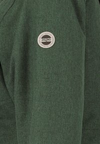 Veste en tissu vert avec une finition texturée et un patch en forme de cercle de couleur grise portant le texte "ZIGZAG" sur le côté gauche.