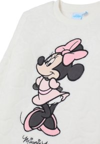 Tela blanca con un bordado de Minnie Mouse vestida con un vestido rosa, un lazo y zapatos, de pie con las manos en las caderas y sonriendo.