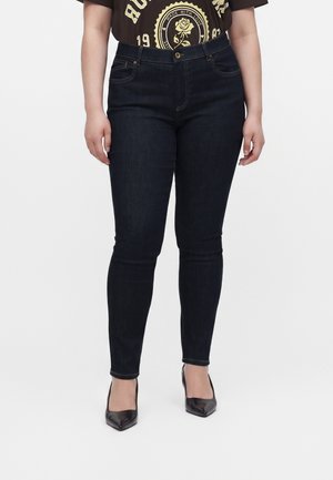 Kvinde iført mørkeblå skinny jeans og sorte spidse hæle, stående mod en ensfarvet hvid baggrund.