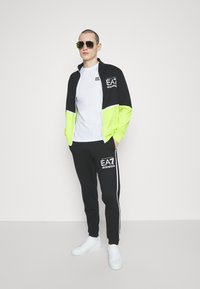 Survêtement noir et vert fluo avec veste zippée et logo, associé à un t-shirt blanc et un pantalon noir avec des rayures latérales, porté avec des lunettes de soleil.
