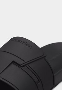 Μαύρο σανδάλι slide Calvin Klein με φαρδιά λουρί που διαθέτει γεωμετρικό κοψίμο και ανάγλυφο όνομα μάρκας.