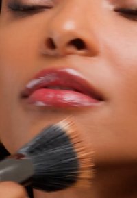 Gros plan d'une personne aux yeux fermés se faisant appliquer du fond de teint sur le menton à l'aide d'un pinceau de maquillage.