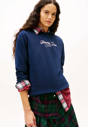 Junge Frau mit langen lockigen Haaren trägt einen marineblauen "Tommy Jeans" Sweatshirt über einem rot-grün karierten Hemd und passendem kariertem Rock.